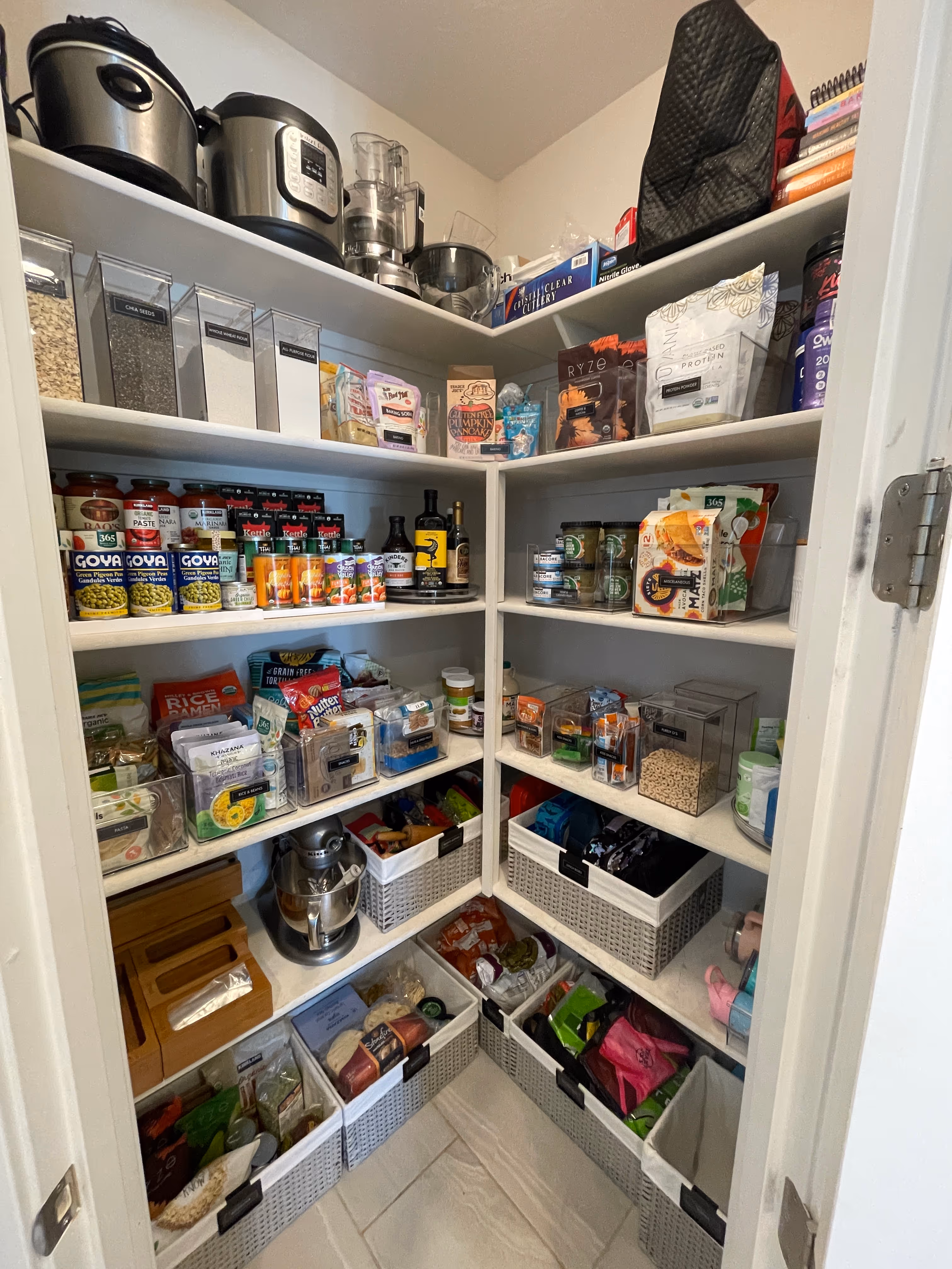 Organized kitchen pantry with shelves holding containers of grains, canned goods, kitchen appliances, dry foods, and baskets with snacks and supplies.