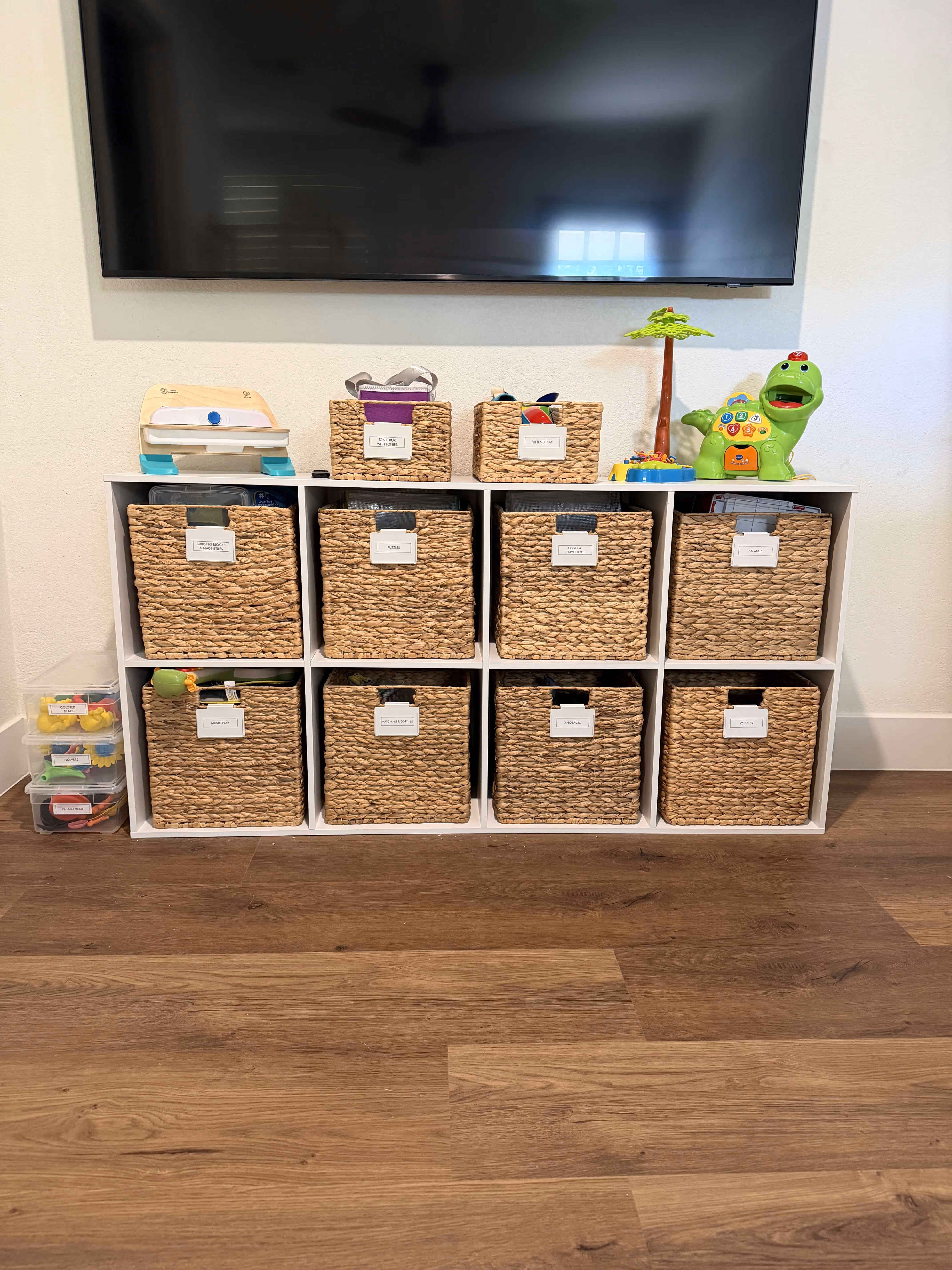 Organized white shelving unit with ten wicker baskets labeled with toy categories below a mounted flat-screen TV, with children's toys on top and plastic containers on the side on wood flooring.