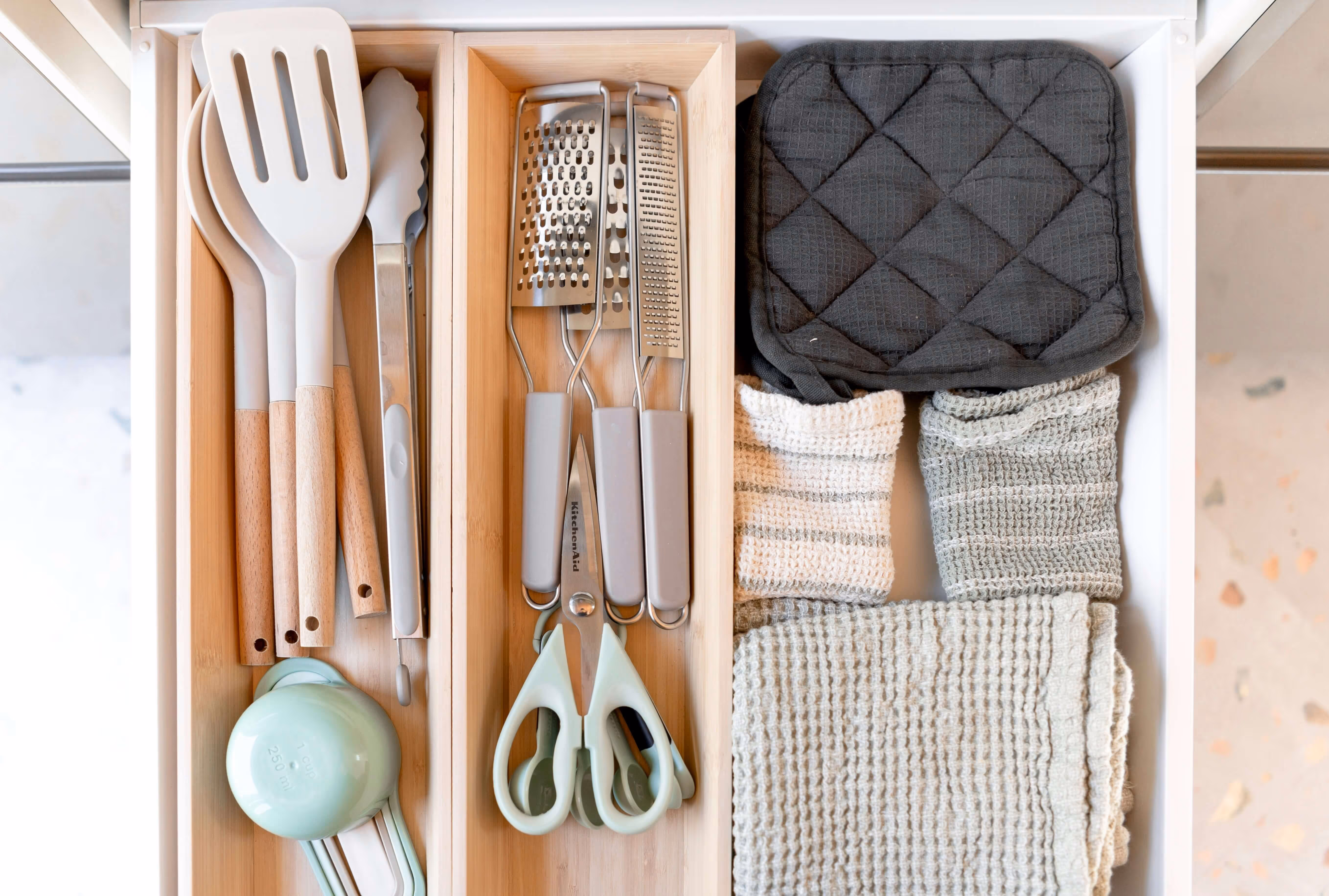 Organized kitchen drawer with wooden spoons, tongs, graters, scissors, measuring cups, black pot holders, and folded kitchen towels.