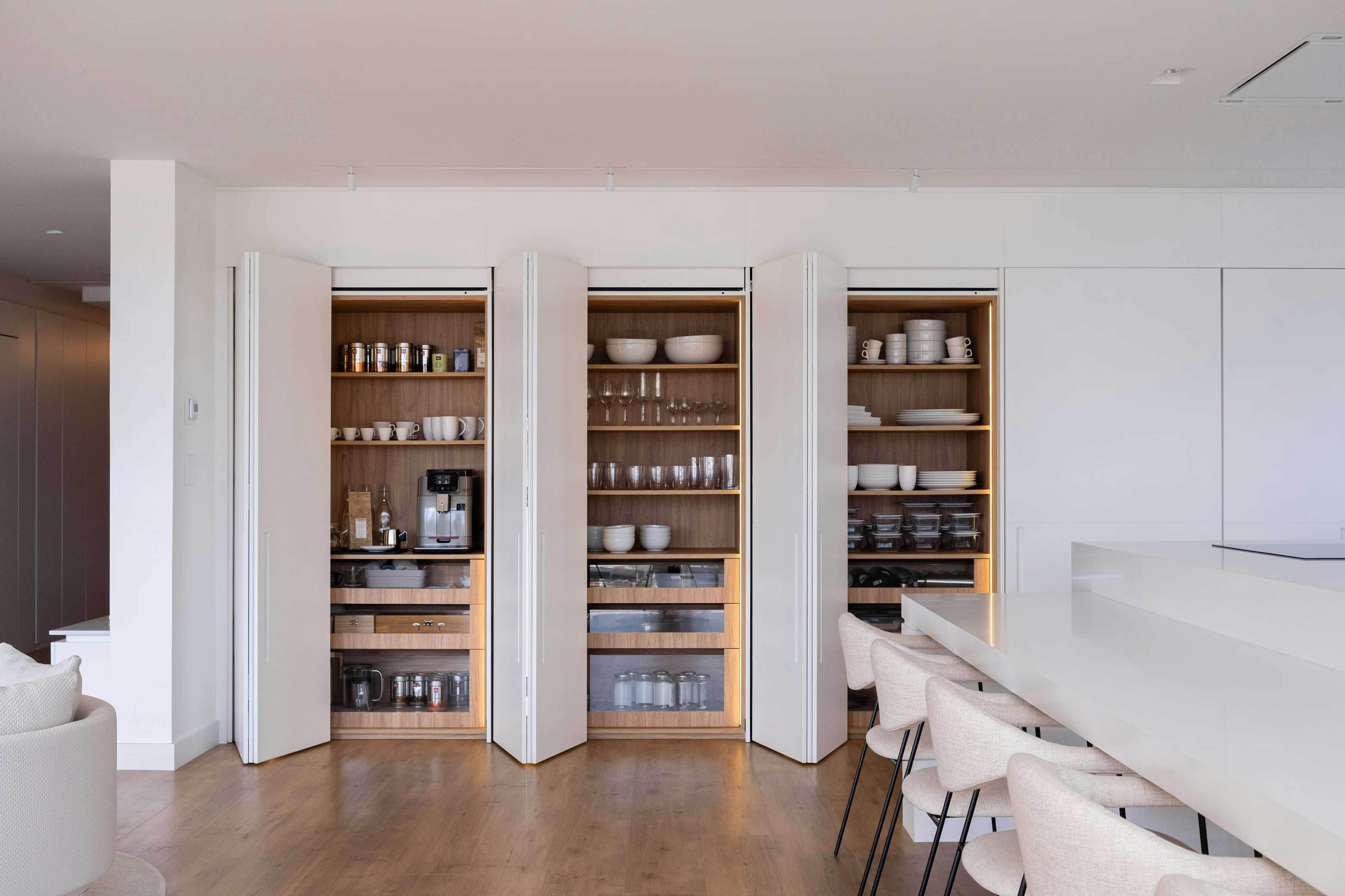 Modern kitchen with three open white pantry cabinets showing shelves with cups, bowls, glasses, coffee machine, and jars.