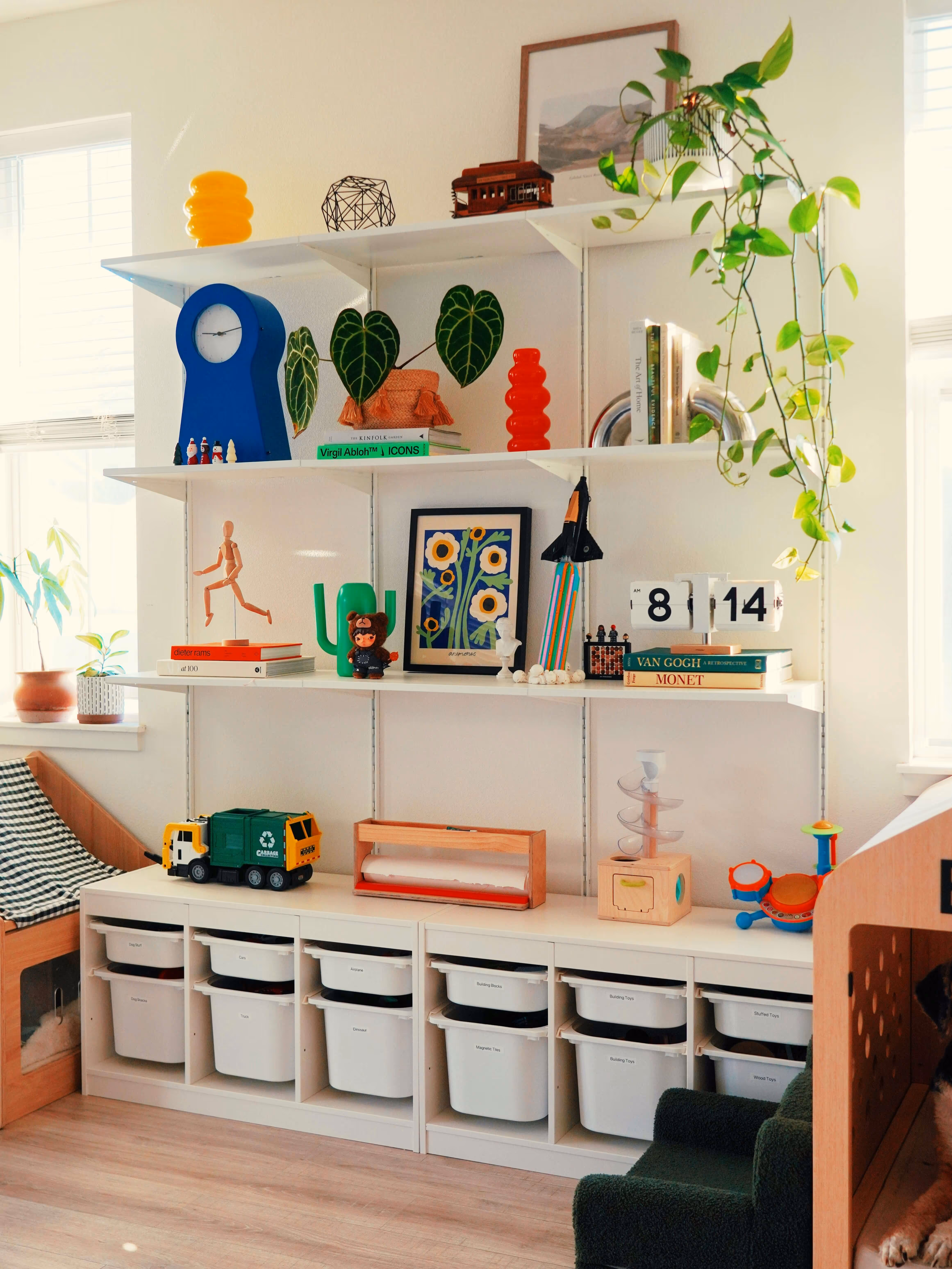 Organized white shelving unit with art, plants, toys, books, and labeled storage bins in a sunlit room.
