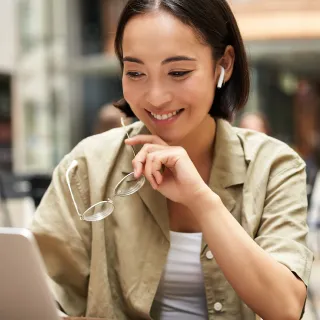 Smiling woman with short hair wearing earbuds and holding glasses, looking at a laptop screen.