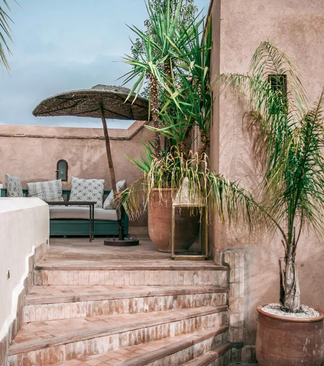 Outdoor seating area with cushioned bench, wicker umbrella, potted palm trees, and terracotta stairs.