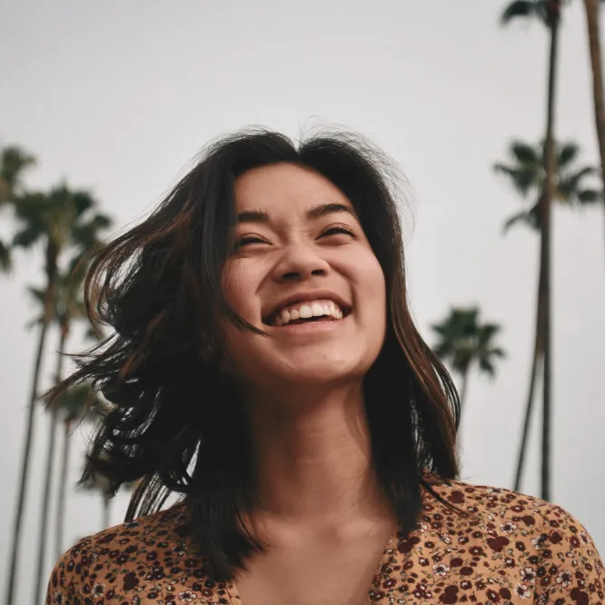 Smiling woman with dark hair outdoors with tall palm trees in the background on a cloudy day.