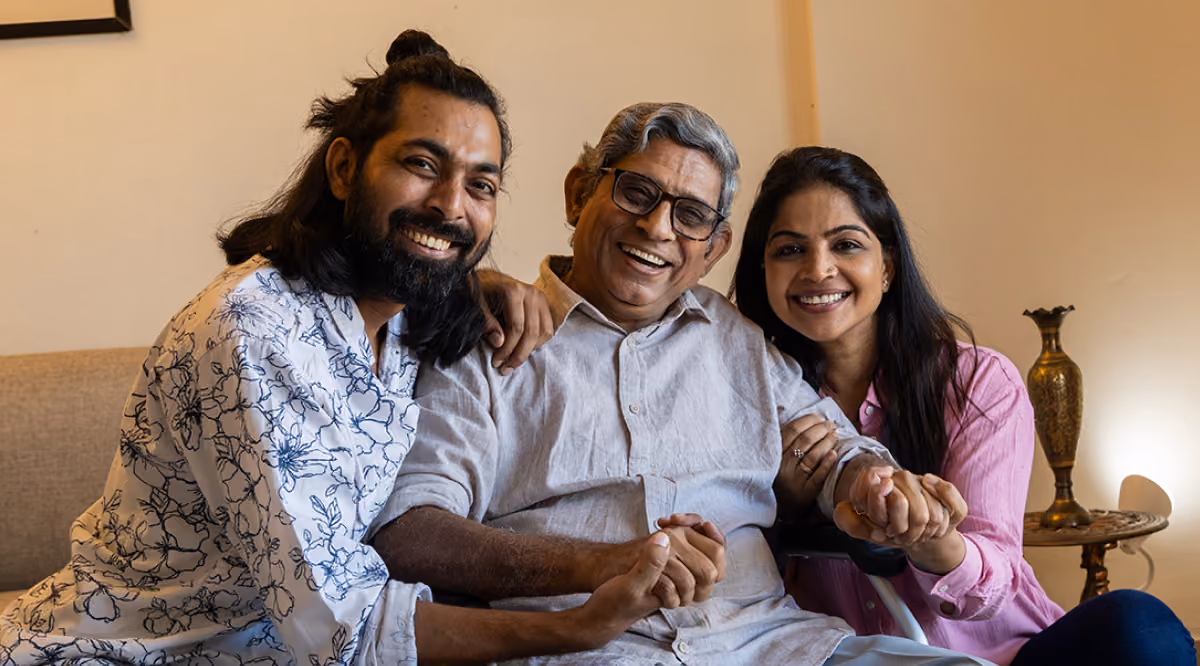 Smiling elderly man with gray hair and glasses hugs two adults, all holding hands on a couch.