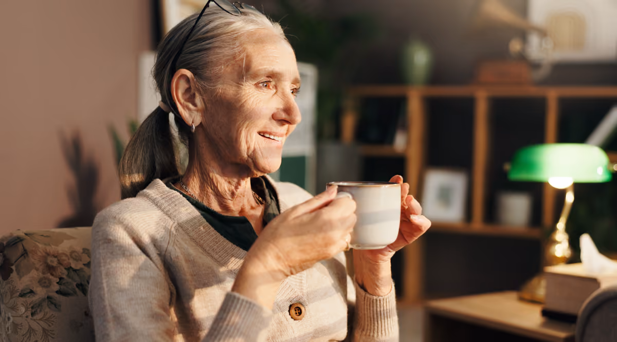 Smiling elderly woman with glasses on head holding a white mug, sitting in a cozy room with lamp and books.
