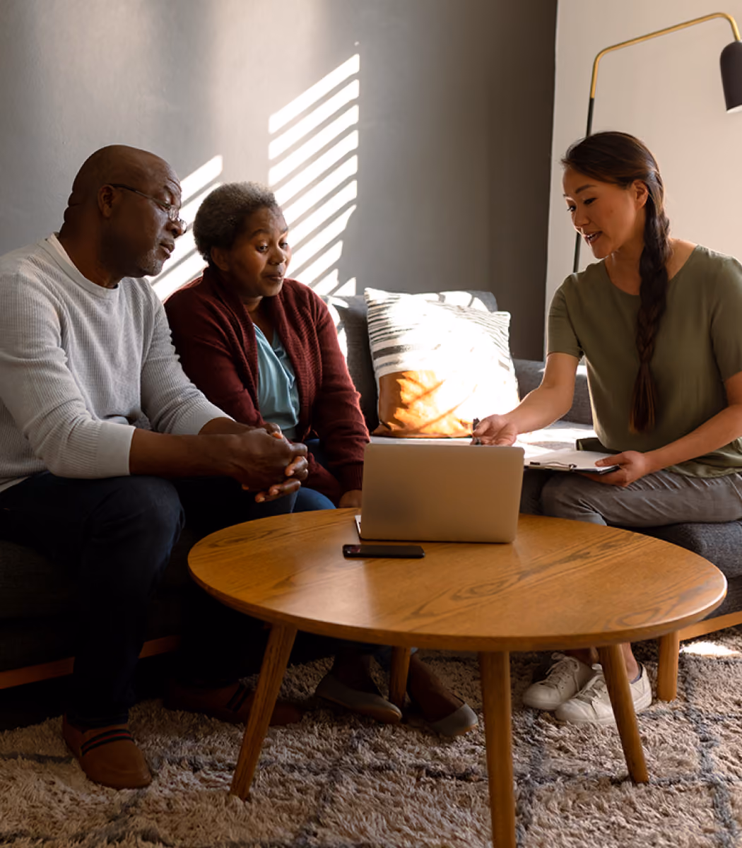 Two older adults sitting on a couch, listening to a woman with a clipboard pointing at a laptop on a table.