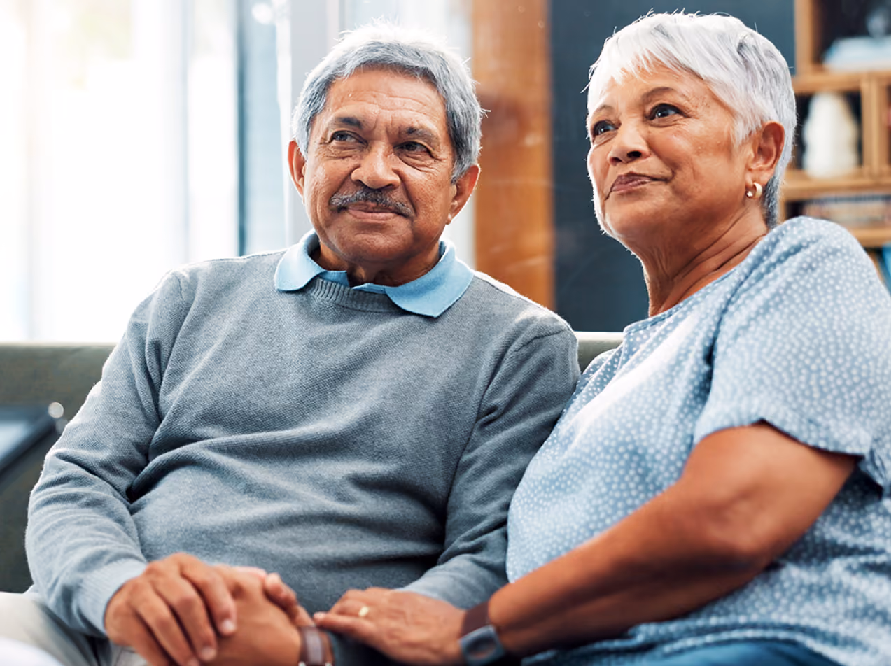 Elderly couple sitting closely on a couch holding hands, appearing content and calm indoors.
