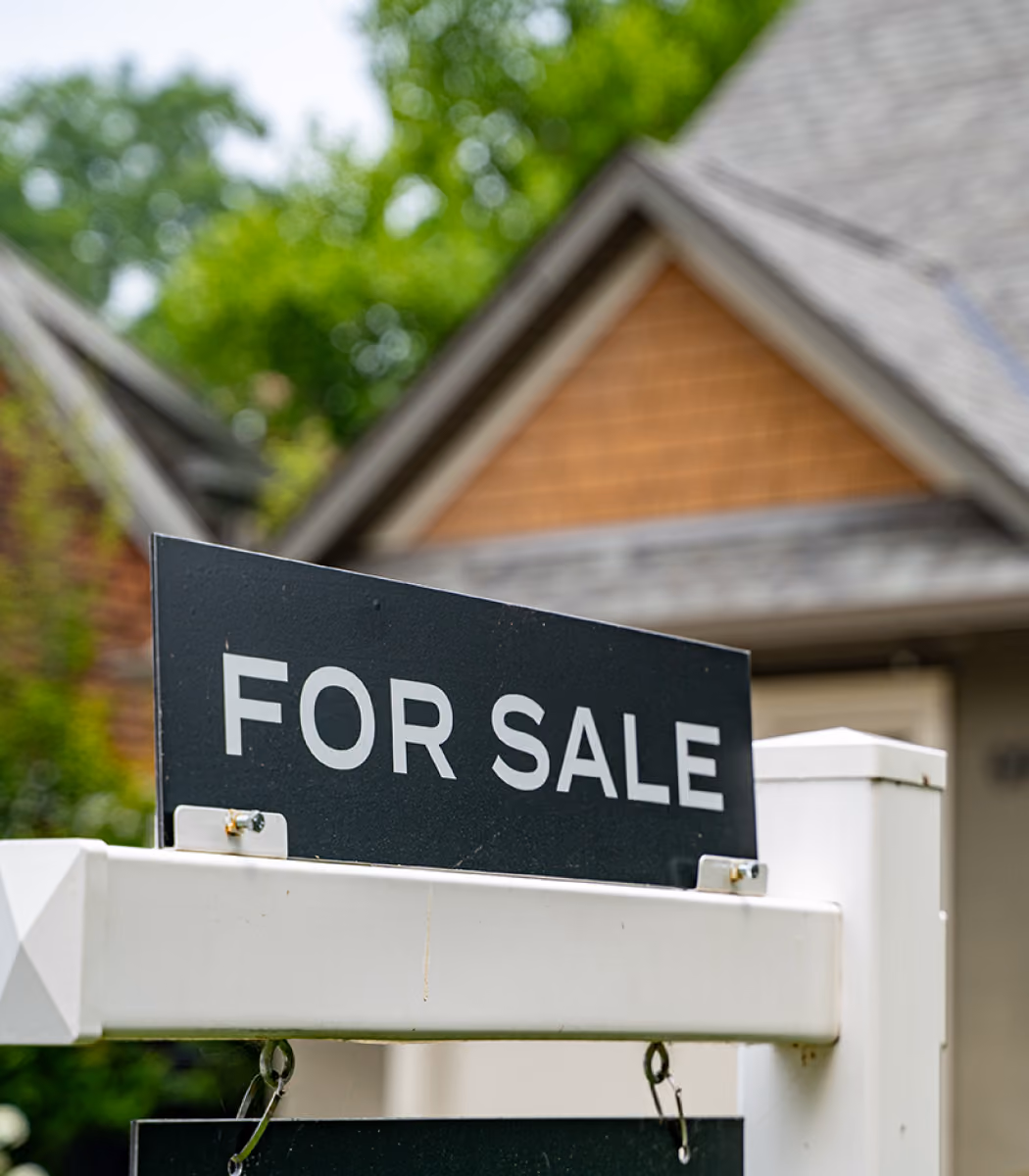 Black and white For Sale sign in front of a house with trees in background.