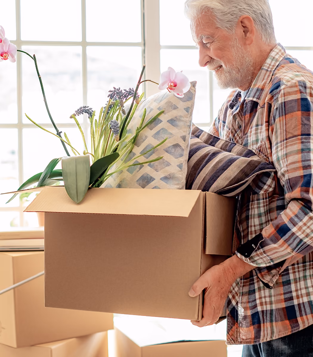 Smiling elderly man holding a box with plants and cushions while moving into a bright room.