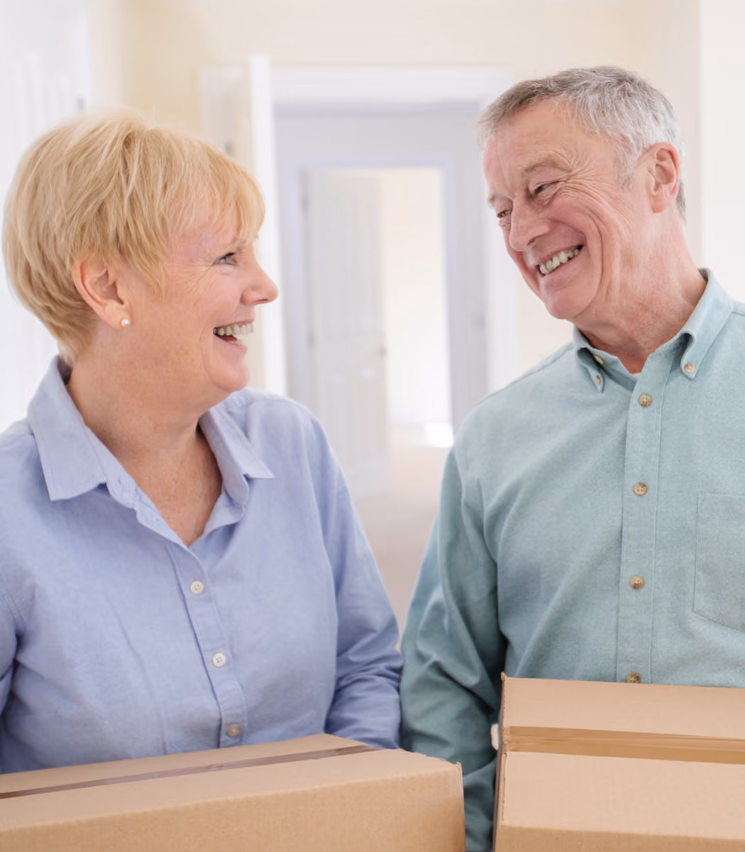 Smiling elderly couple holding moving boxes and looking at each other in a bright room.