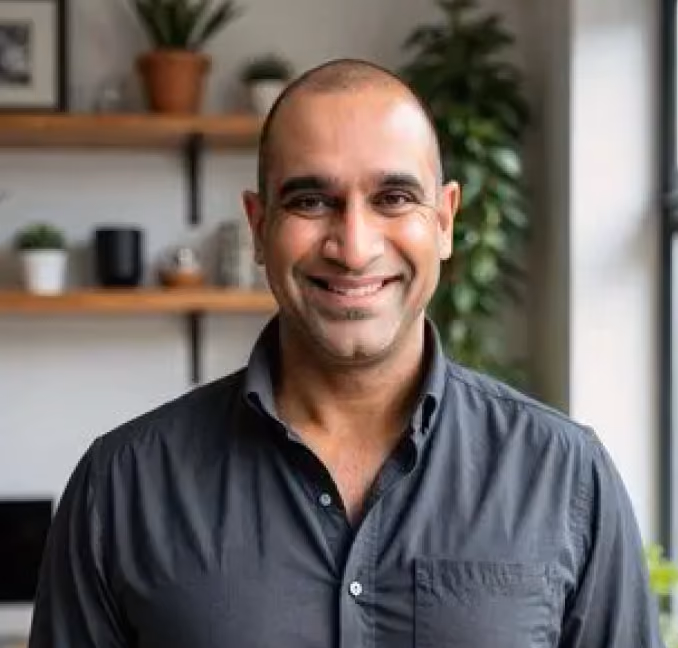 Smiling bald man wearing a dark shirt standing indoors with plants and shelves in the background.