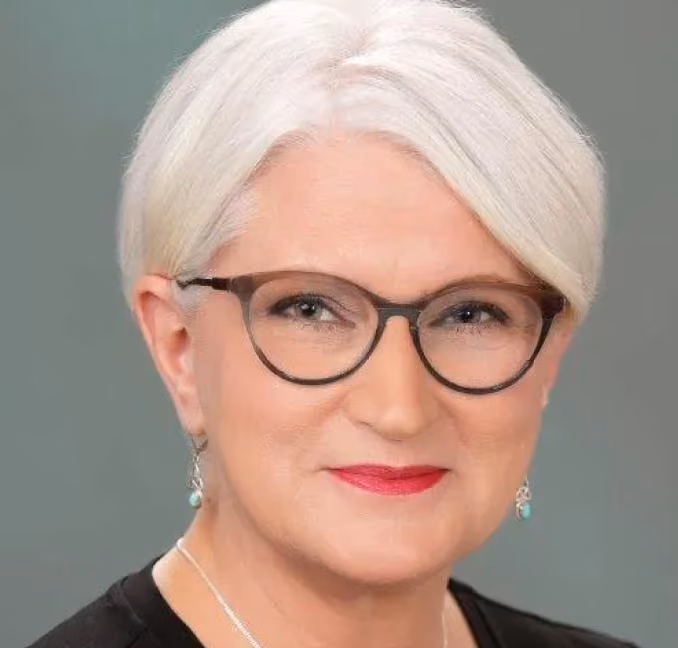 Smiling older woman with short white hair, glasses, red lipstick, and turquoise earrings.