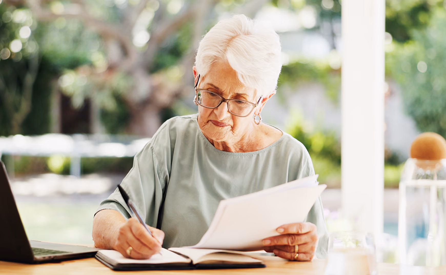 Elderly woman with glasses writing in notebook while holding papers at a table with a laptop outside.
