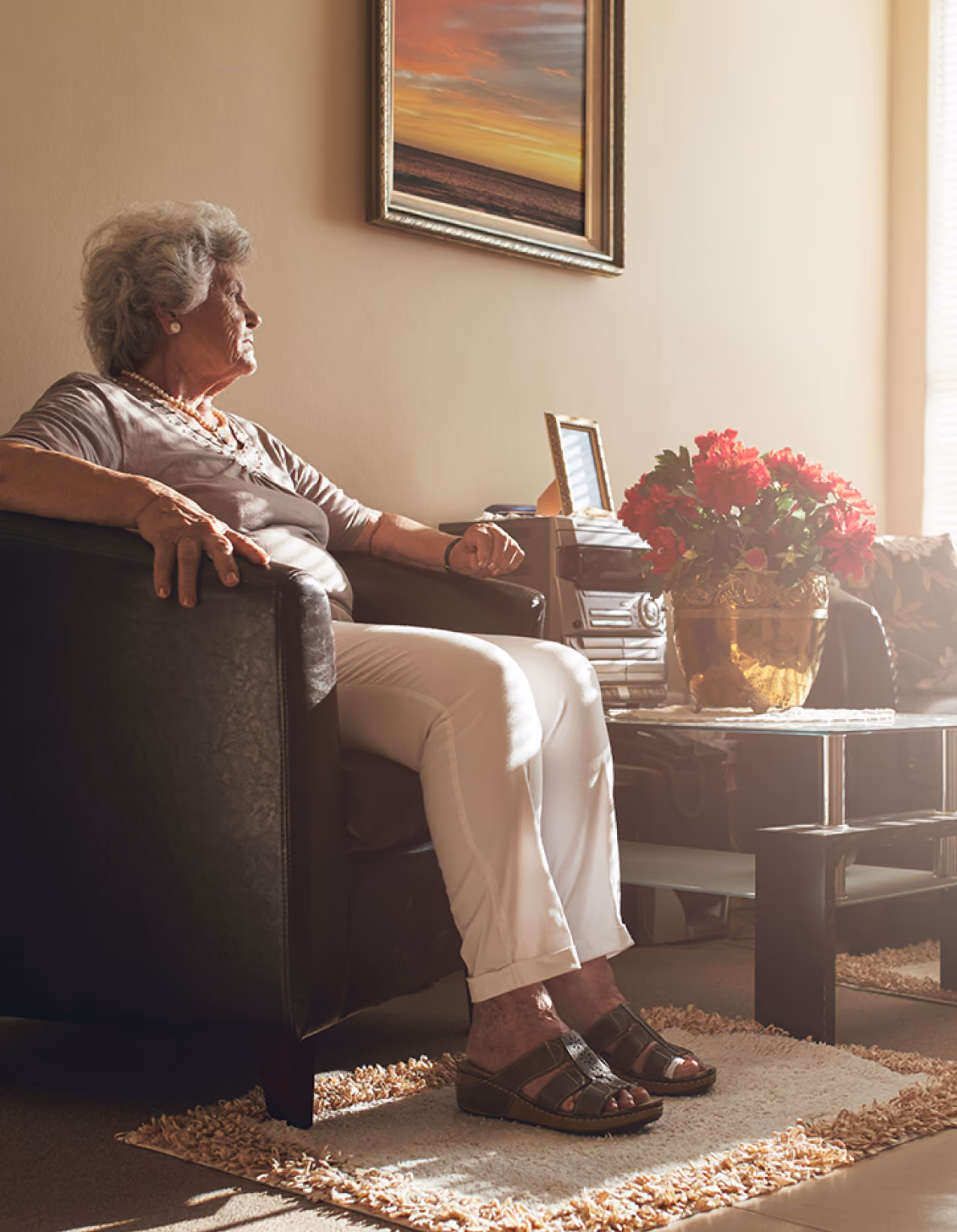 Elderly woman sitting in a black chair in a sunlit room next to a table with red flowers.