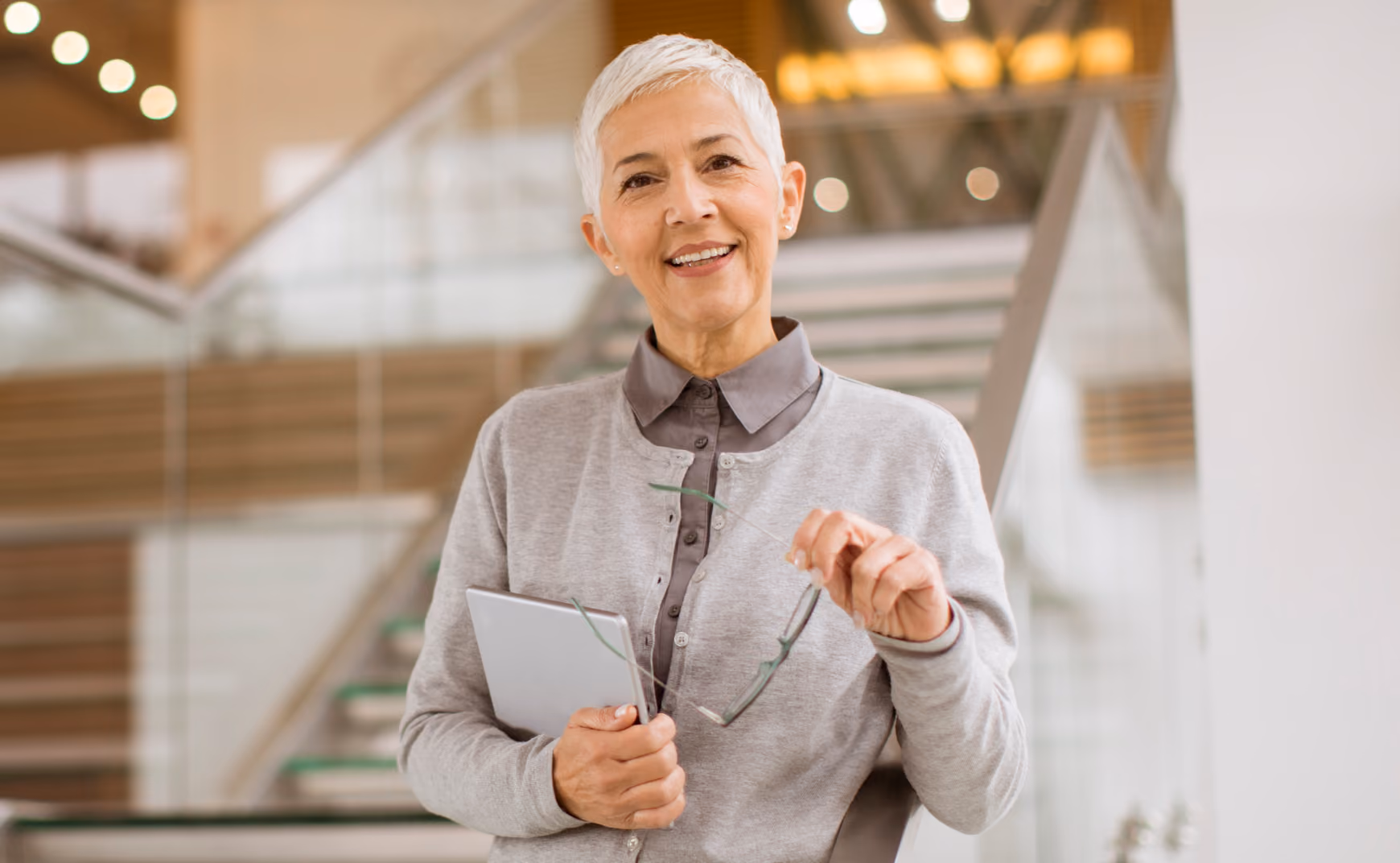Smiling older woman with short white hair holding glasses and a tablet in a modern building.
