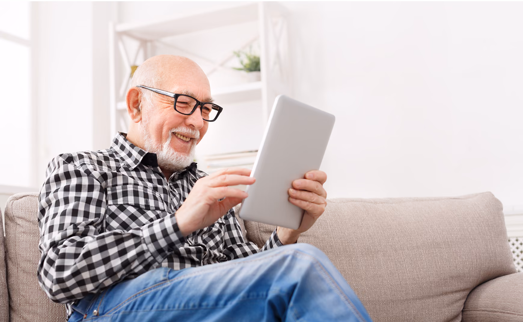 Smiling elderly man wearing glasses and checkered shirt using a tablet while sitting on a beige sofa.