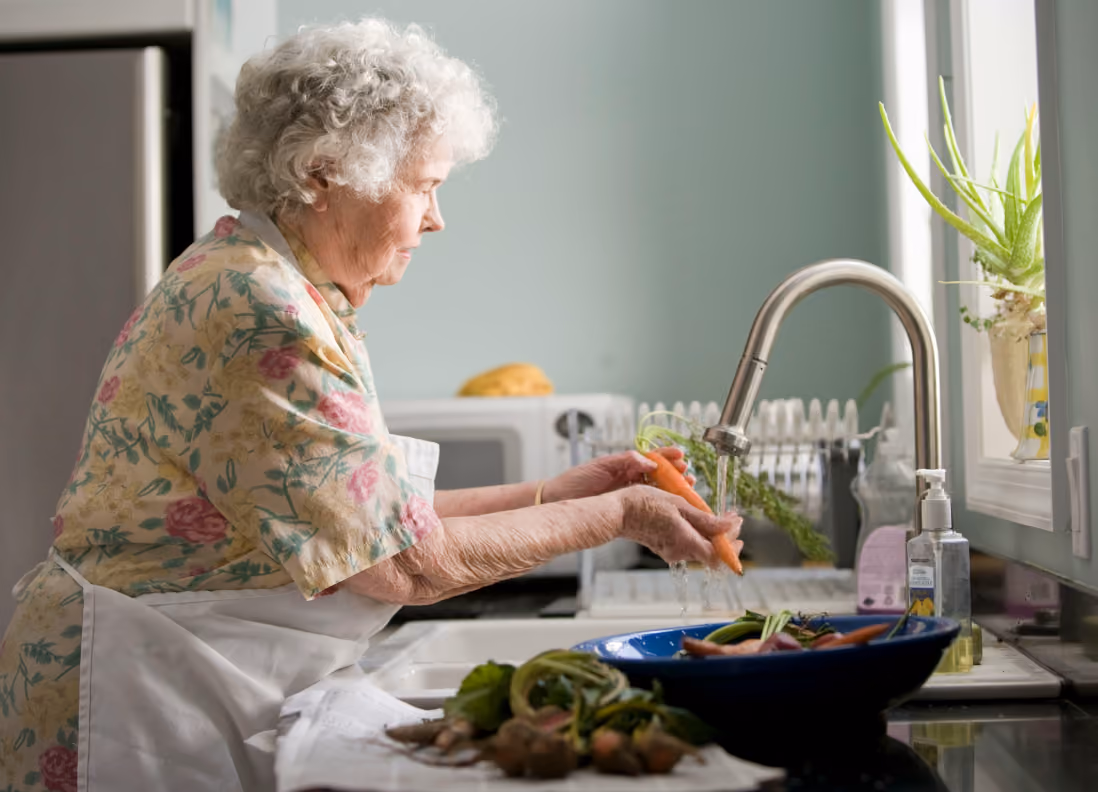 Elderly woman washing carrots under kitchen faucet in a home kitchen.
