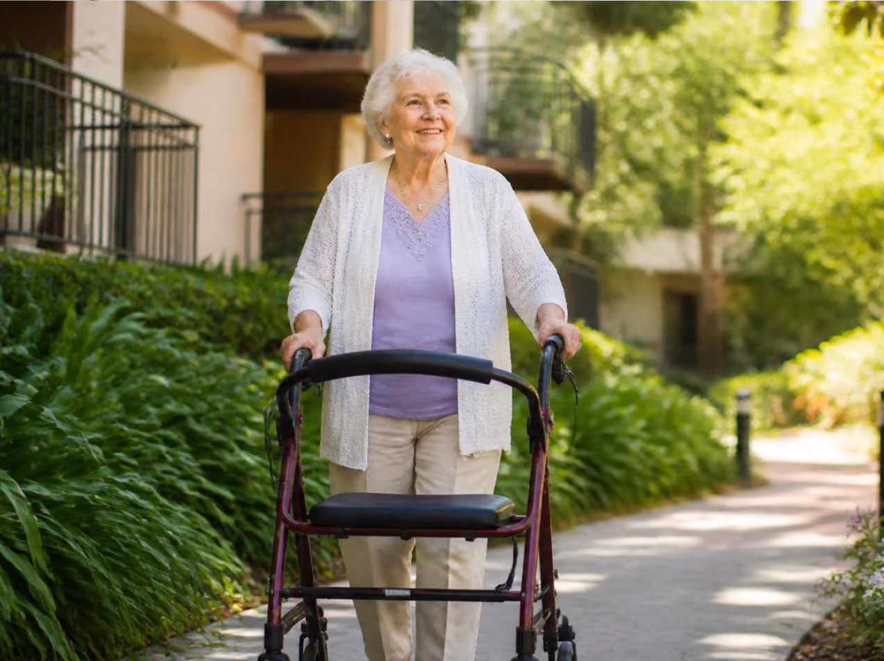 Elderly woman walking outside on a path using a walker, surrounded by green plants and buildings.