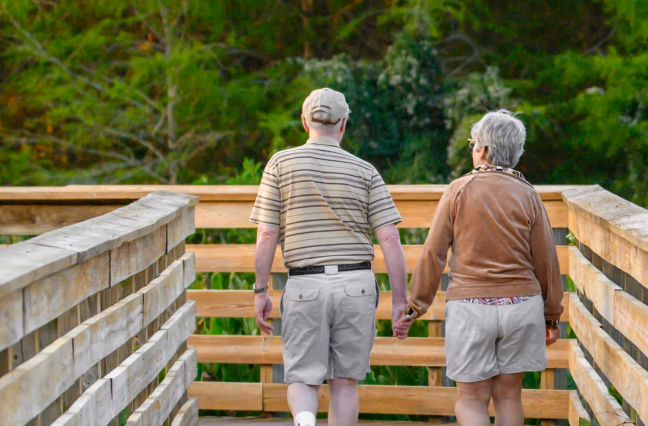 An elderly couple holding hands walking on a wooden bridge surrounded by green trees.