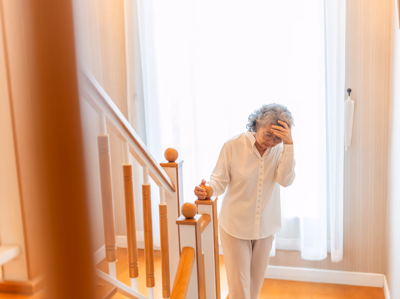 Elderly woman holding head and staircase railing, showing signs of dizziness or discomfort indoors.