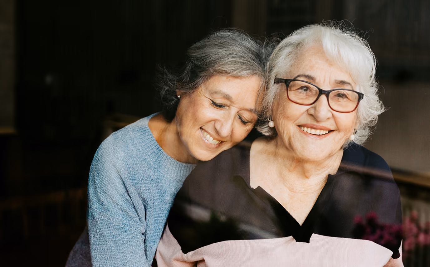 Older woman with glasses smiling as a middle-aged woman hugs her from behind with eyes closed.