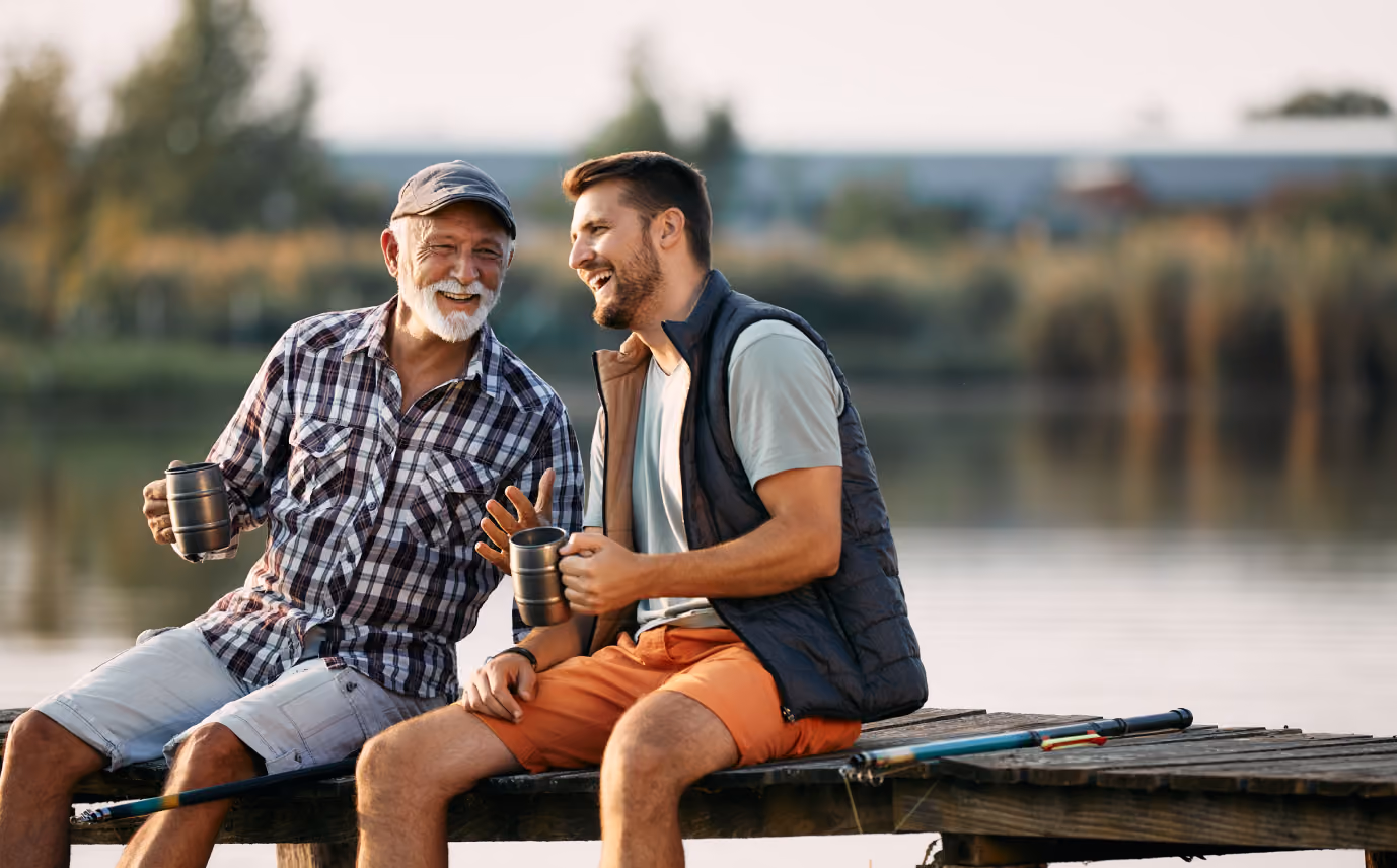 Older and younger men sitting on dock by lake, holding mugs and laughing with fishing rods nearby.