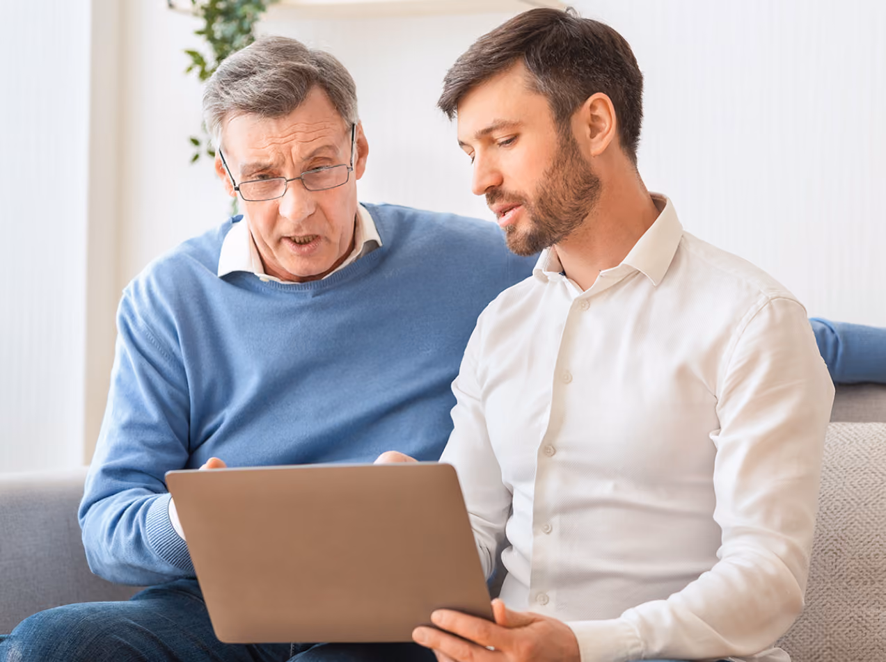 Two men, one older with glasses and one younger with a beard, looking at a laptop together.