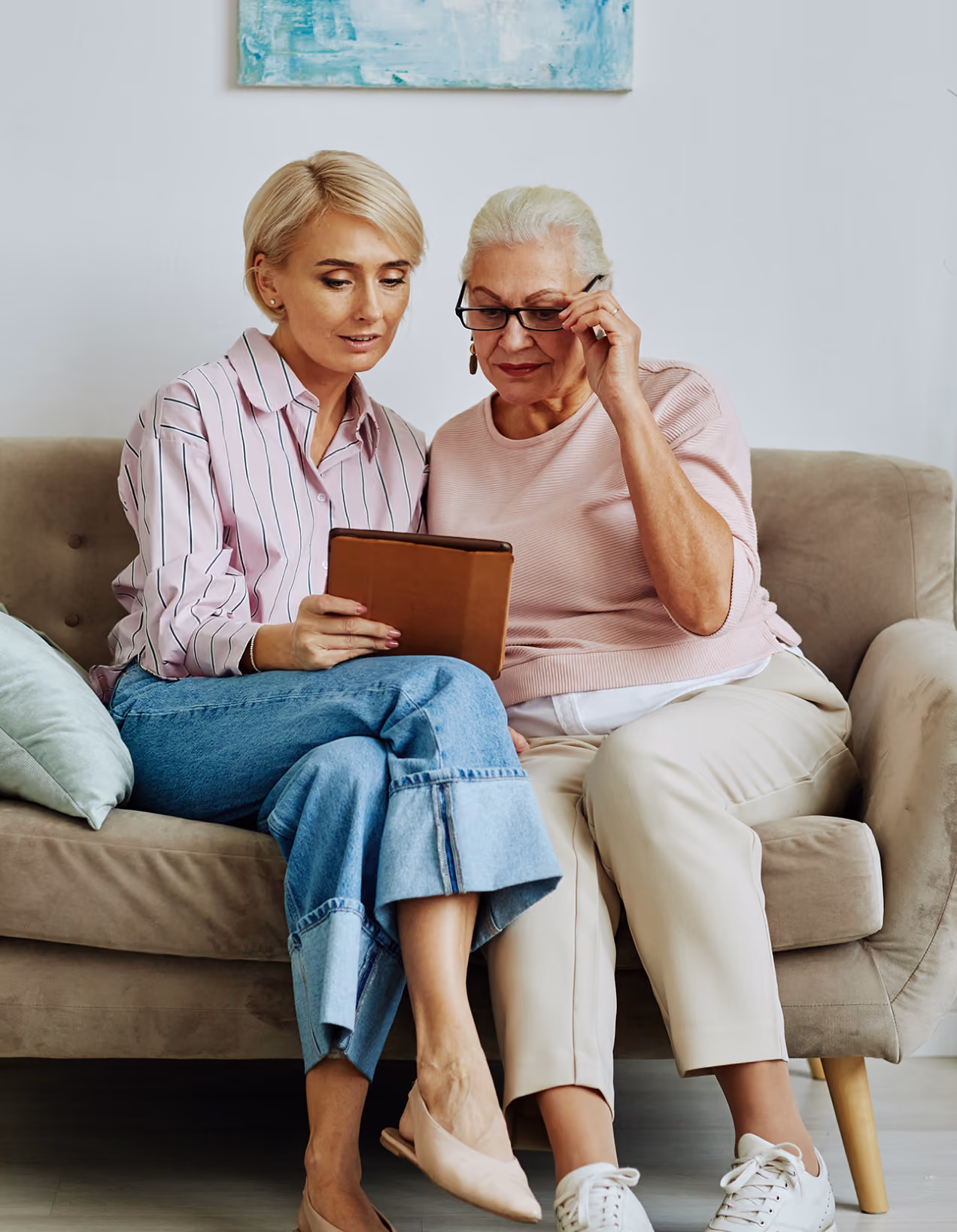 Two women sitting on a couch looking at a tablet, one adjusting her glasses.