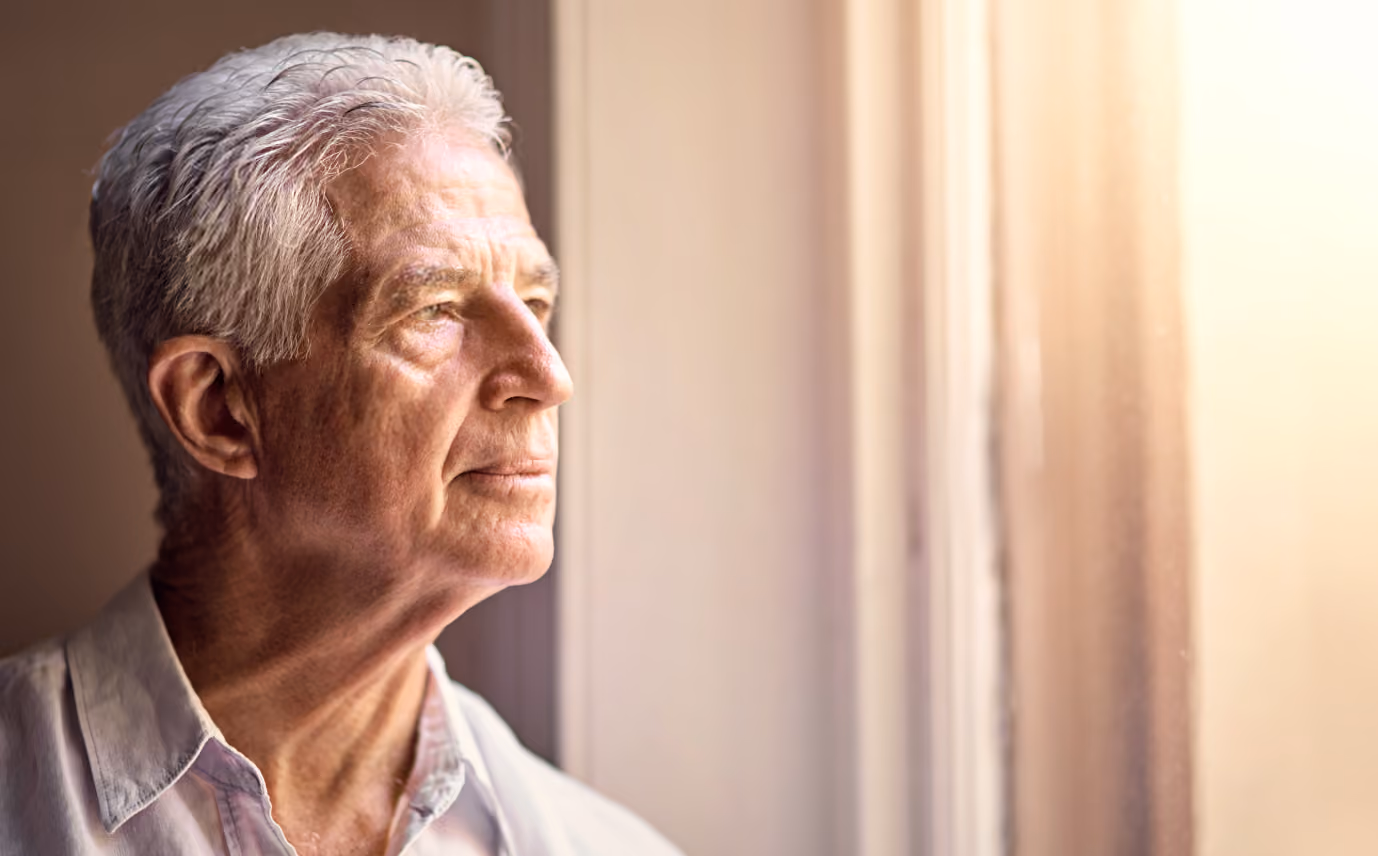 Elderly man with gray hair gazing thoughtfully out a sunlit window.
