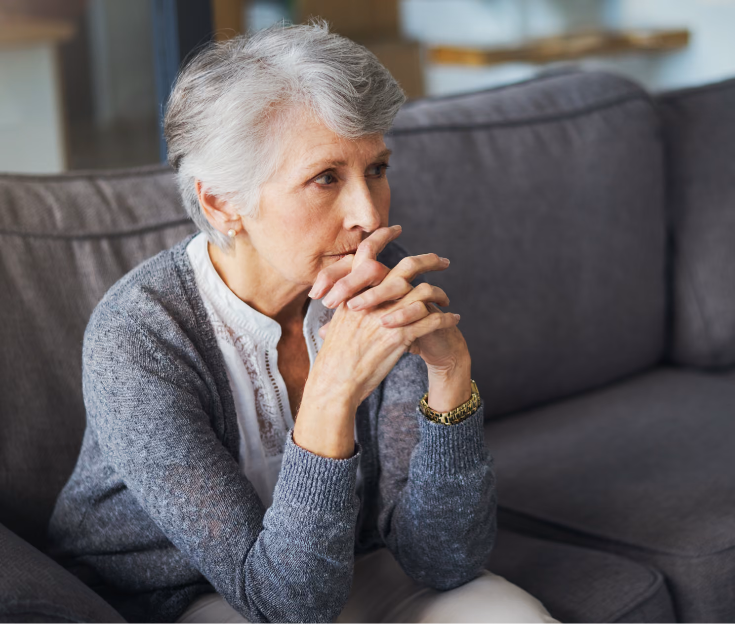 Older woman sitting on a couch, looking thoughtful with hands clasped near her mouth.