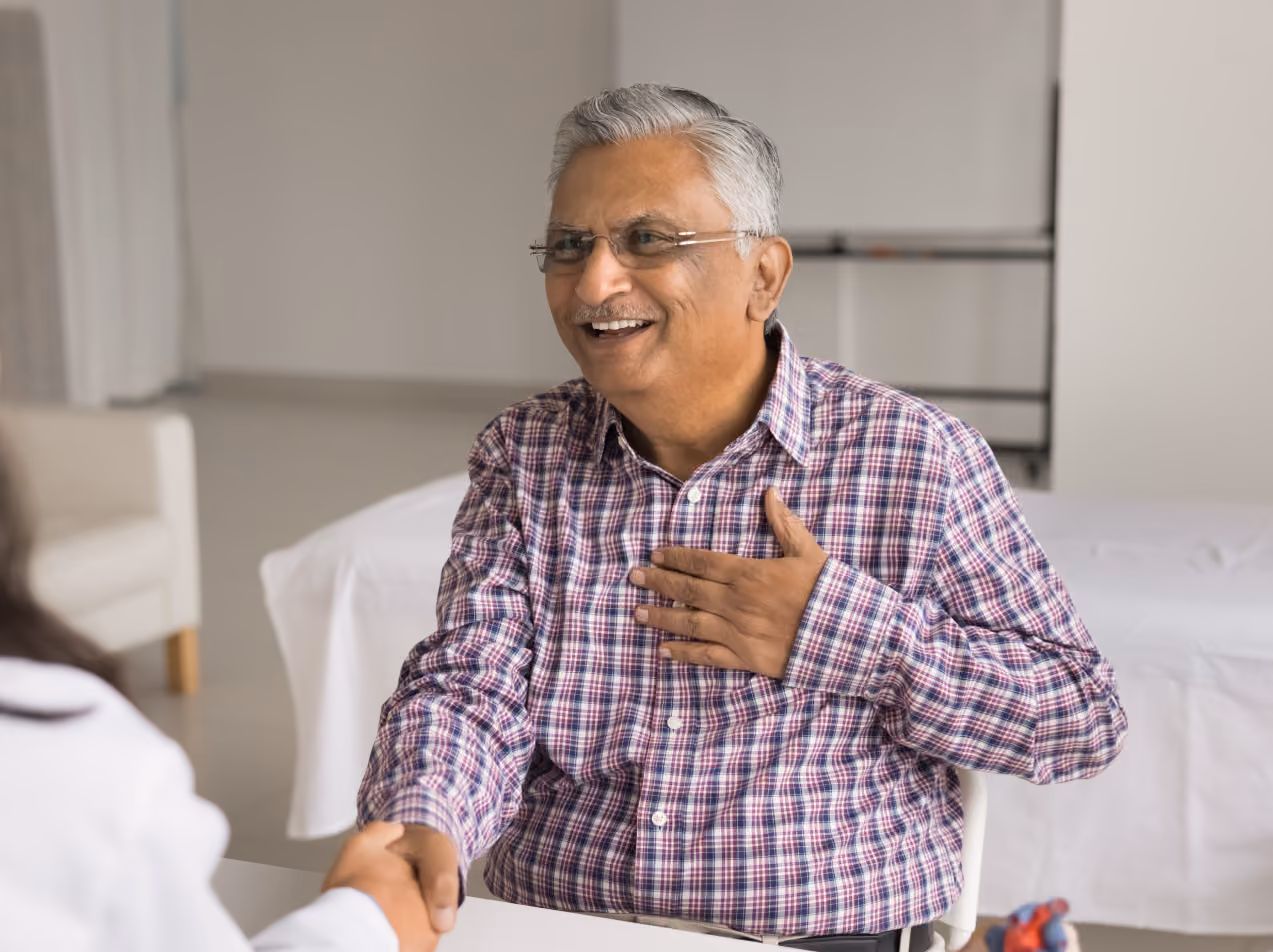 Smiling elderly man in glasses shaking hands and placing his hand on his chest in a bright room.