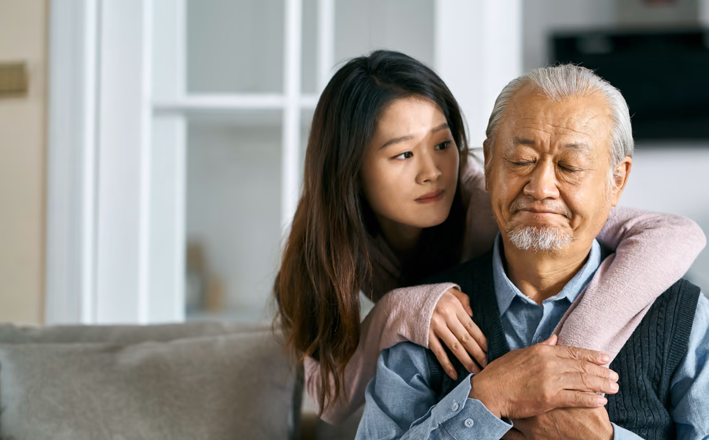 Young woman comforts elderly man sitting with closed eyes and hands clasped on chest.
