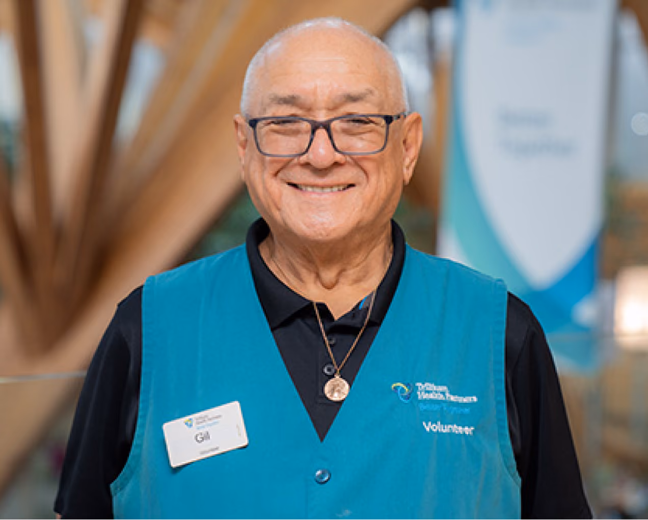 Smiling elderly man wearing glasses and a teal volunteer vest with a name tag reading Gala.