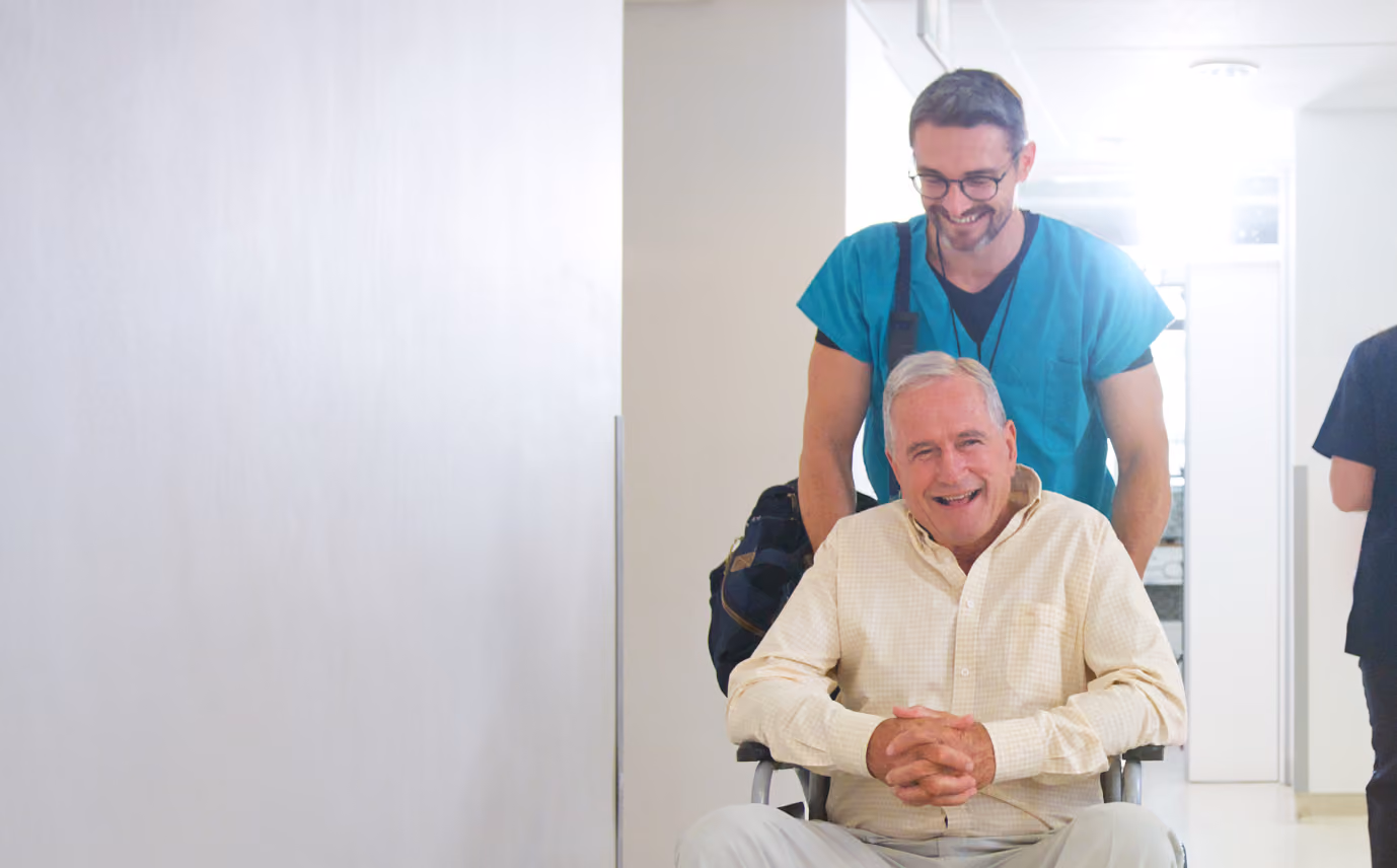 Smiling senior man in wheelchair pushed by a smiling healthcare worker in scrubs down a bright hallway.