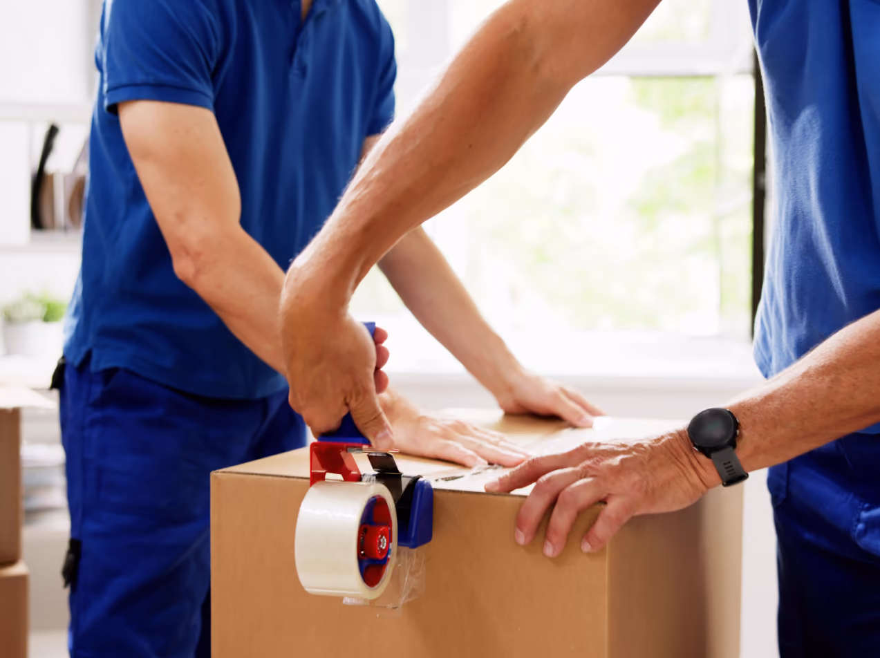 Two men in blue shirts sealing a cardboard box with packing tape indoors.