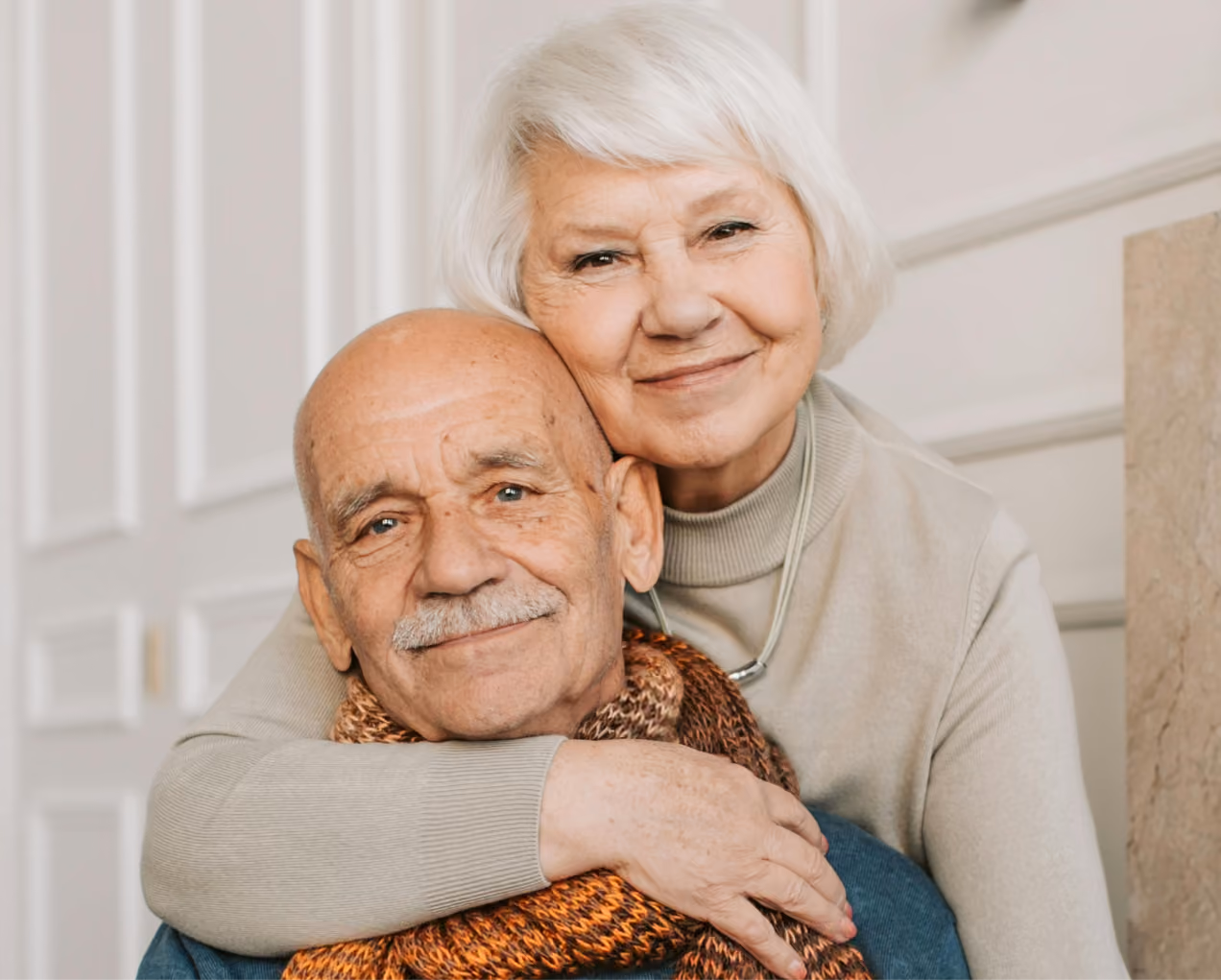 Smiling elderly woman with white hair hugging a bald elderly man wearing a scarf indoors.