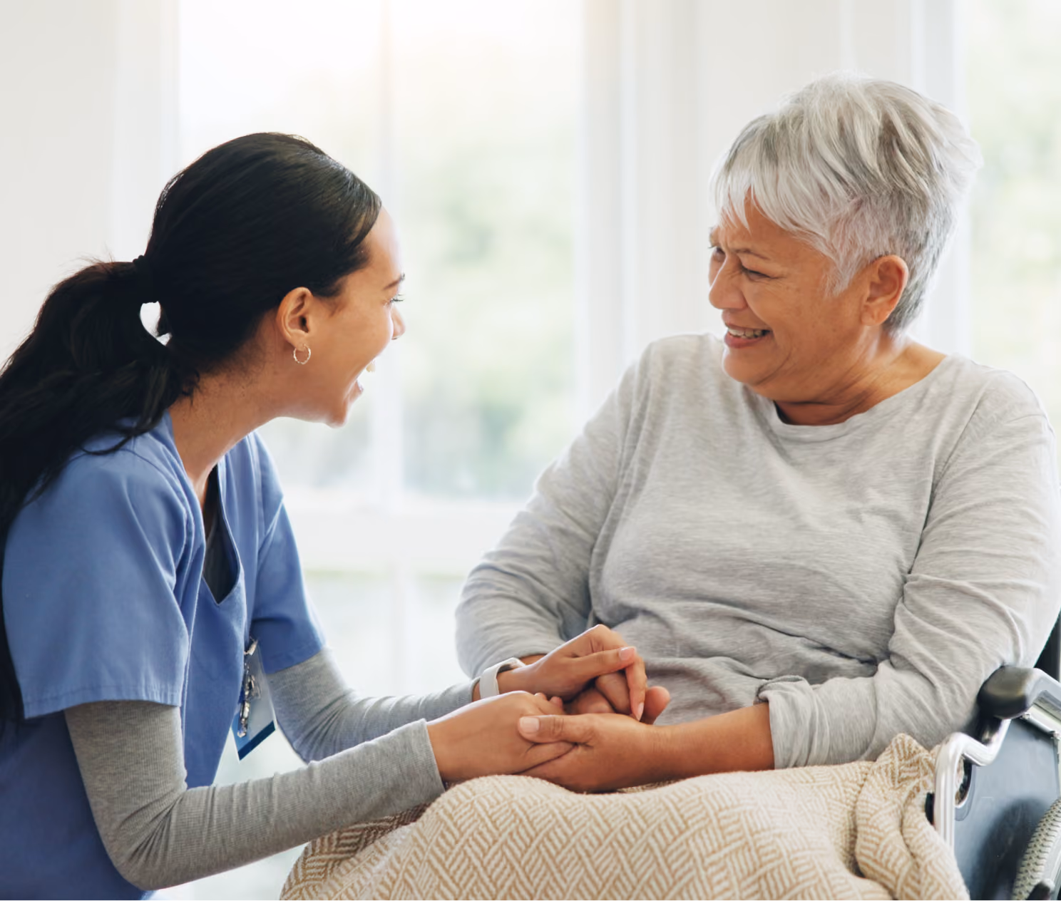 A healthcare worker in scrubs holding hands and smiling with a senior woman in a wheelchair.