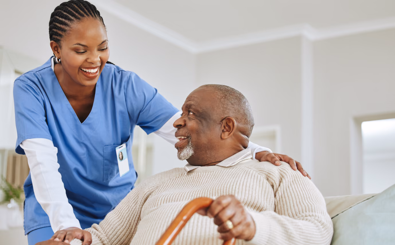 Smiling nurse in blue scrubs comforting elderly man in beige sweater holding a wooden cane.