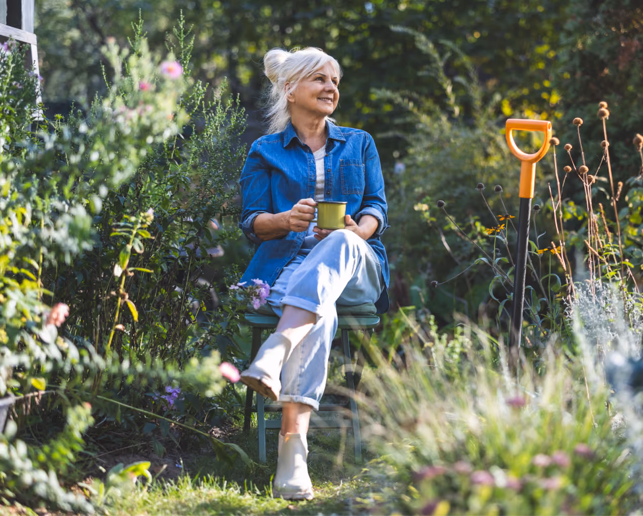 Smiling older woman sitting in garden with cup, surrounded by plants and garden tools.