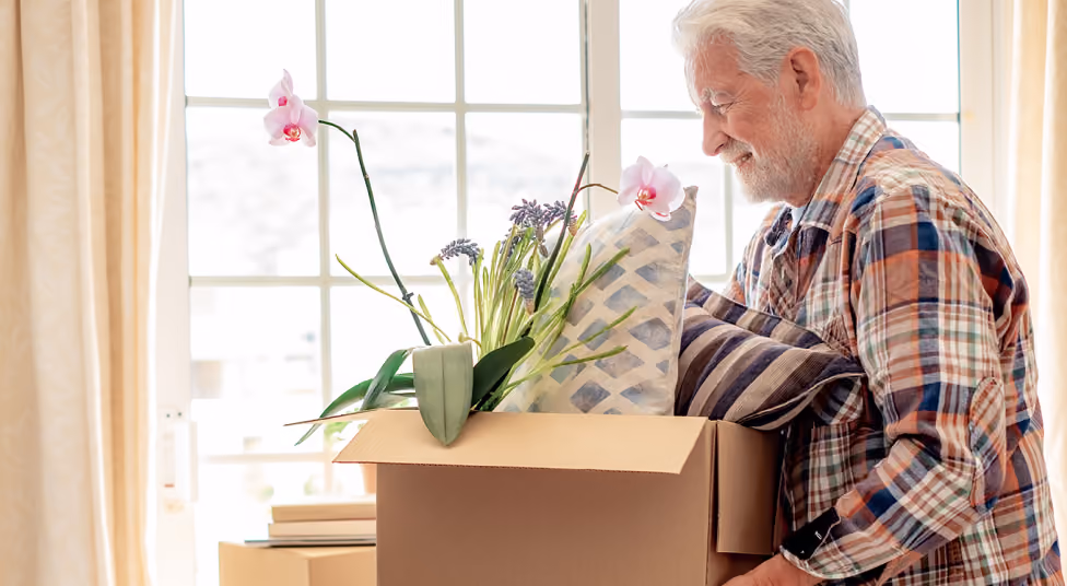 Elderly man holding cardboard box with pillows and flowers in bright room near window.