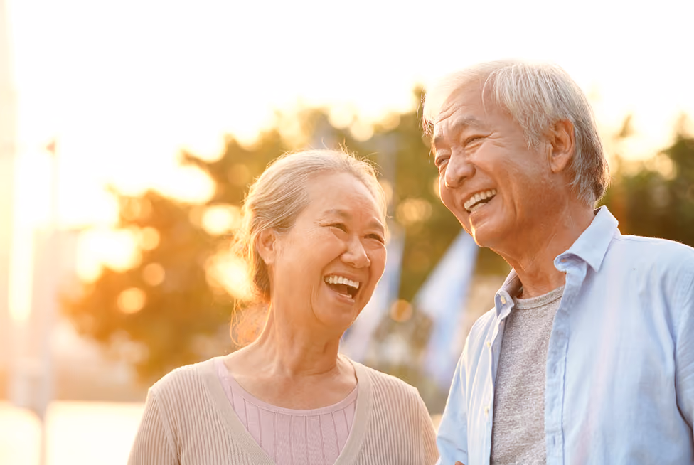 Smiling elderly Asian couple outdoors enjoying a sunny day with blurred trees in background.