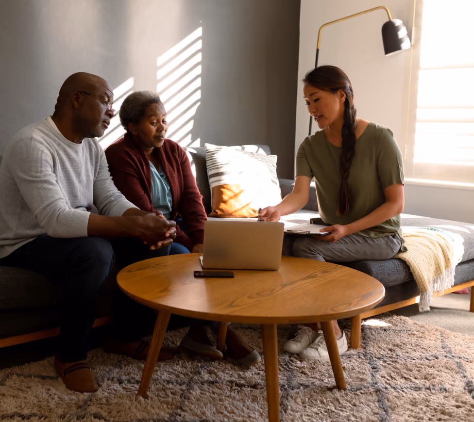 A woman with a clipboard shows a laptop screen to an older couple sitting on a couch in a sunlit room.
