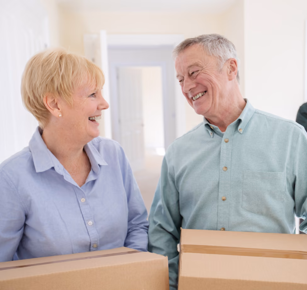 Smiling elderly couple holding cardboard boxes inside a bright, empty home.