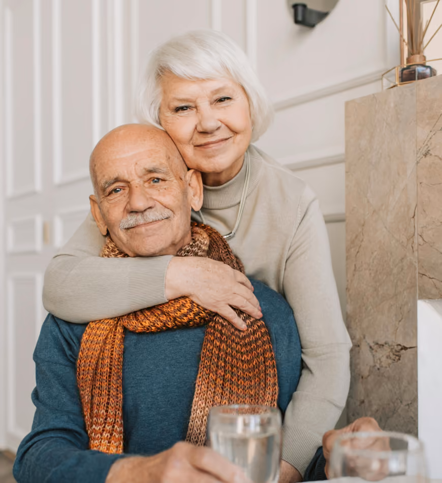 Elderly woman hugging smiling elderly man who wears a scarf while seated indoors.