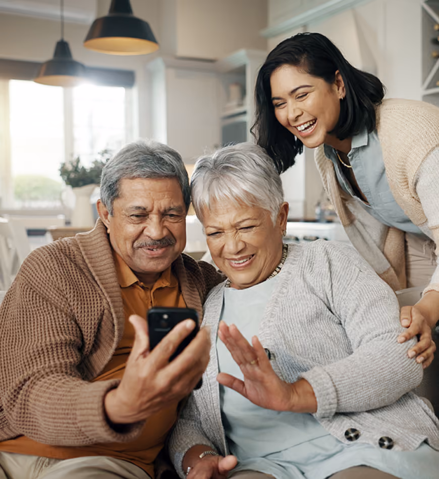 Three people smiling and looking at a smartphone in a cozy home setting.