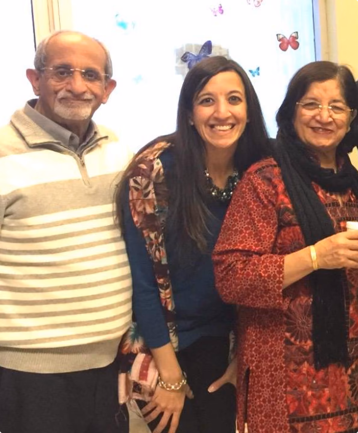 Three people smiling: an older man in a striped sweater, a woman with long hair, and a woman in red patterned attire.