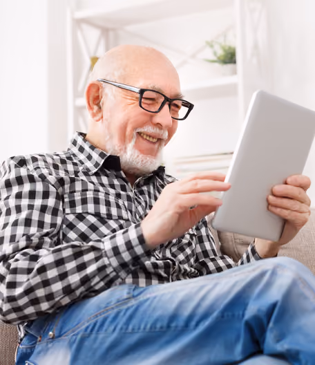 Smiling elderly man wearing glasses using a tablet while sitting on a couch at home.