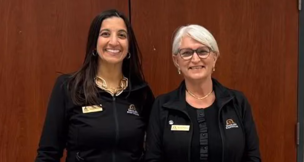 Two women standing and smiling in black jackets with name tags in front of a wooden door.