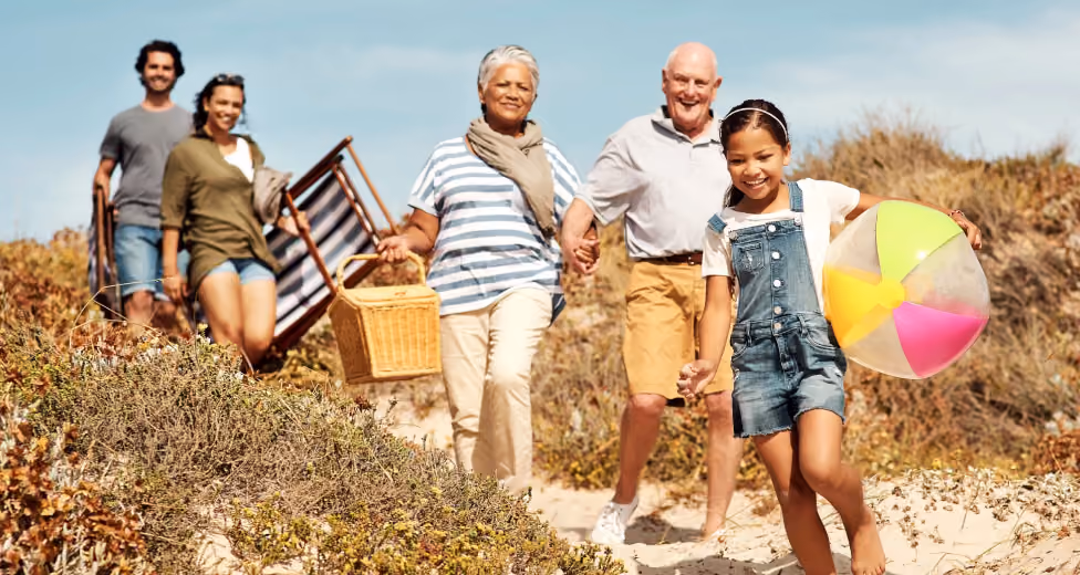 Multigenerational family walking on a sandy beach path carrying a picnic basket, chair, and beach ball.